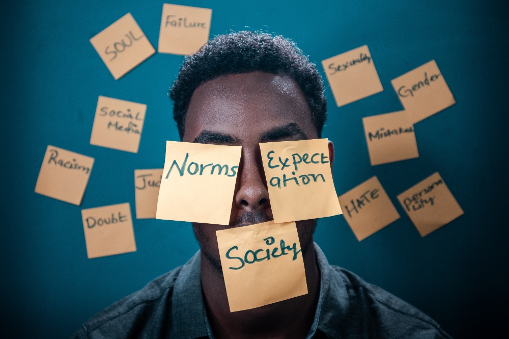 Black man with post-it notes on his face, covering is eyes and mouth, and around his head with words like "norms," "society," and "expectations.""